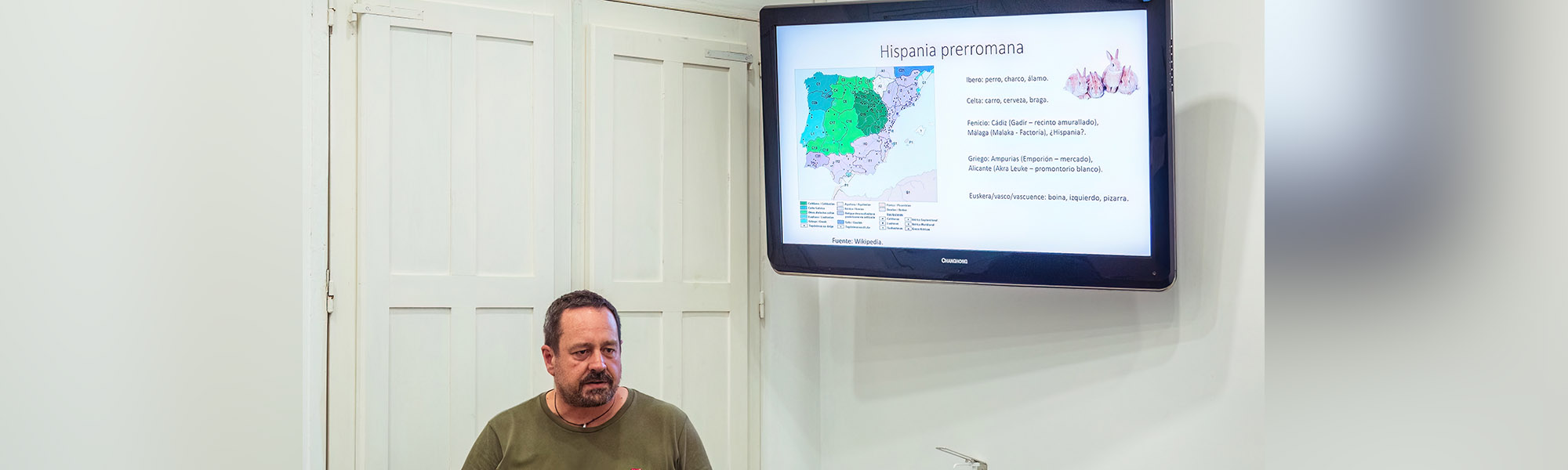 A person giving a lecture with a digital screen displaying a slide at the back